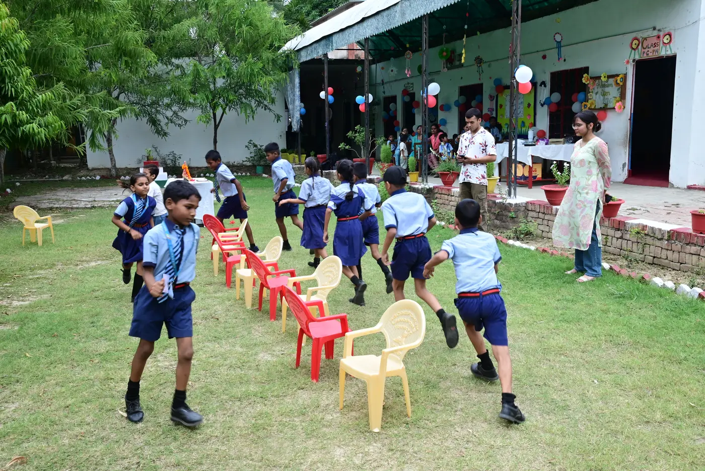 Children playing musical chairs on the lawn at Lords Public School, running around a row of red and yellow chairs.