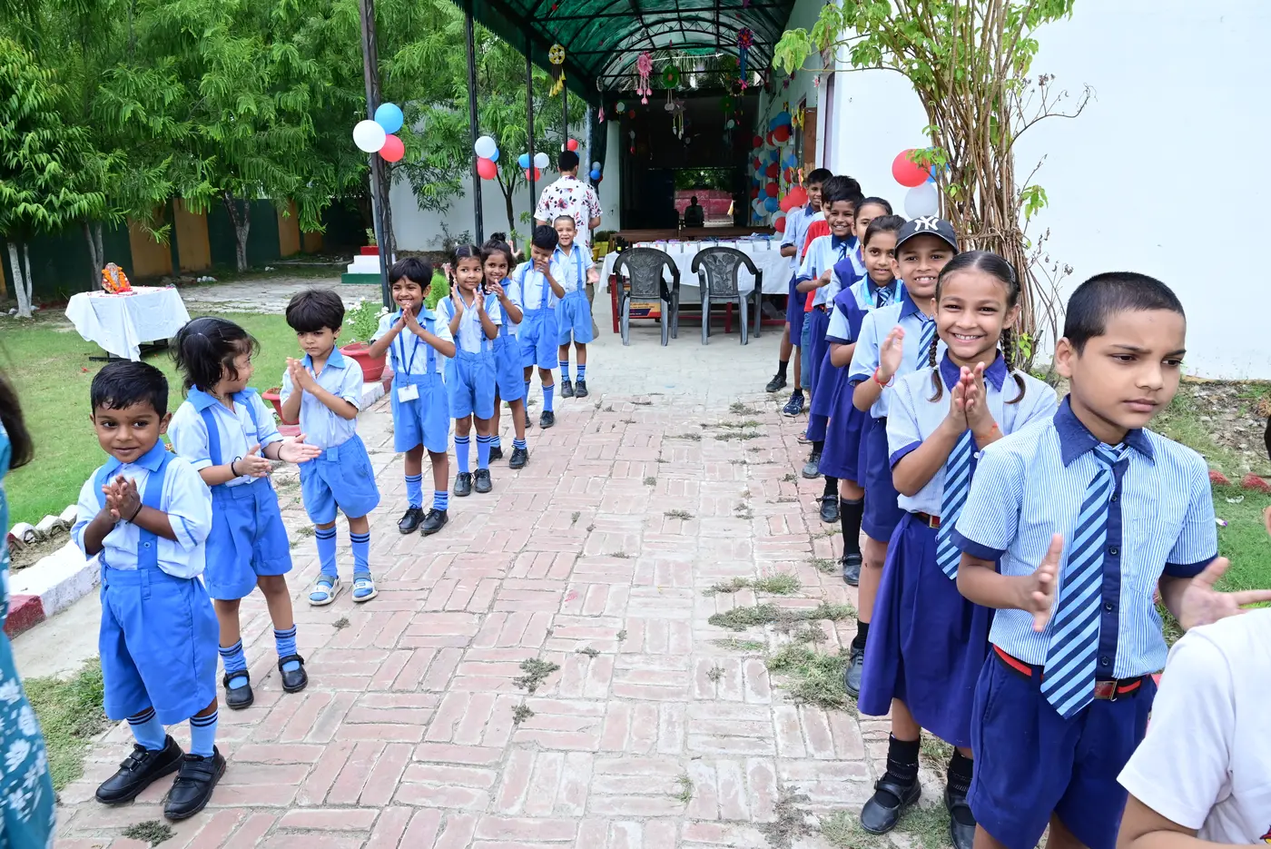 Children of Lords Public School standing along the brick path in their blue uniforms, clapping and smiling as they welcome visitors.