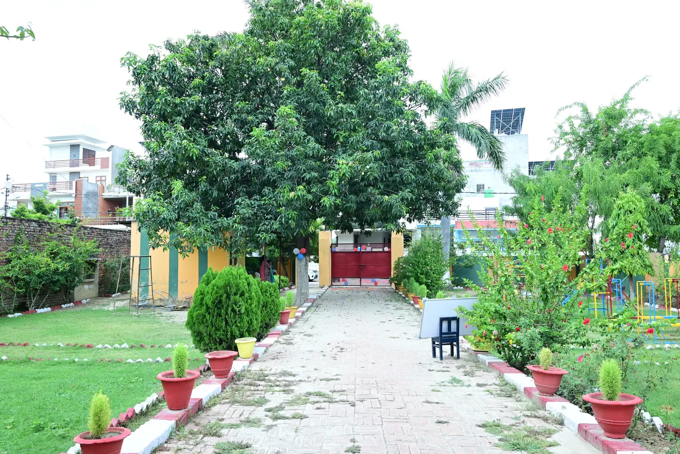 The brick path leading from the entry gate into the Lords Public School campus, lined with potted plants and shaded by a large mango tree.