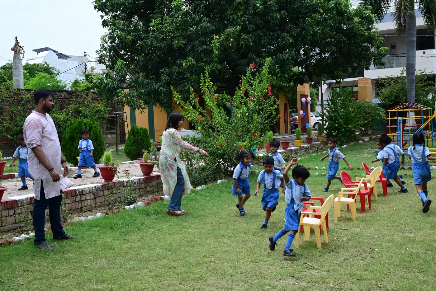 A teacher at Lords Public School starting a game on the lawn while children run past a row of chairs.